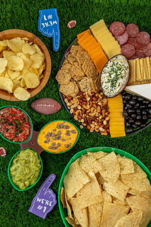 Display of bowls of chips, platter of cheese, crackers, olives, nuts, salami and nuts with spinach articoke dip, salsa, cheese dip and guacamole and football tailgate themed party decorations portrait orientationの写真素材