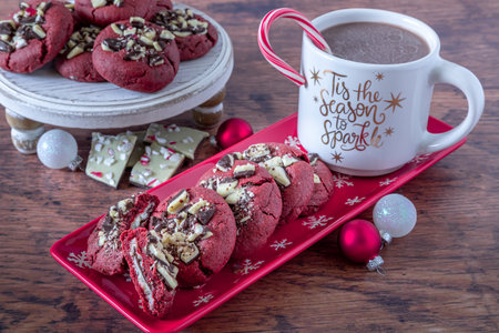 Holiday baking display of peppermint bark stuffed red velvet cookies on a red plate with snowflakes, a cup of hot chocolate with a candy on rim and plate of cookies in backgroundの写真素材