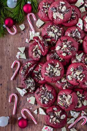 Overhead shot of homemade peppermint bark stuff red velvet cookies stacked on wooden table surrounded by peppermint bark candy pieces and small candy canes, with evergreen branches and ornament decorationsの写真素材