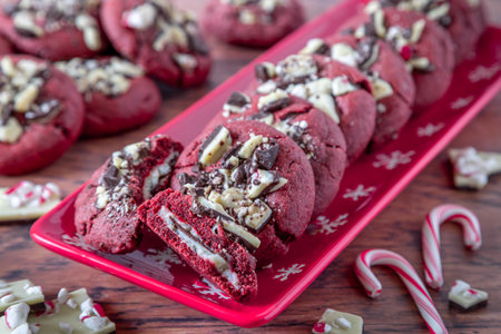 Close up of cookie display of peppermint bark stuffed red velvet cookies on a red plate with snowflakes, and stack of extra cookies in the background, candy canes in front and pieces of peppermint bark candyの写真素材