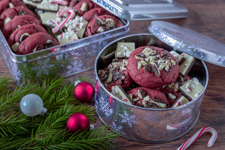 Food gift of peppermint bark stuffed red velvet cookies in a silver cookie tins with snowflakes, small candy canes on wooden table and peppermint bark pieces included with cookies and evergreen branches and ornament decorationsの写真素材