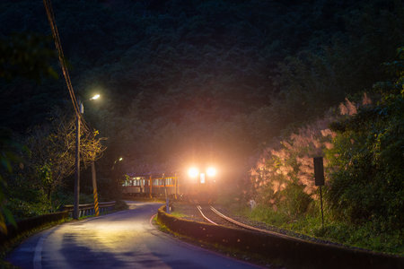 A yellow diesel train passes through the village at night with its lights on. Along the Pingxi line, there are river valleys, potholes and waterfalls. Taiwanの写真素材