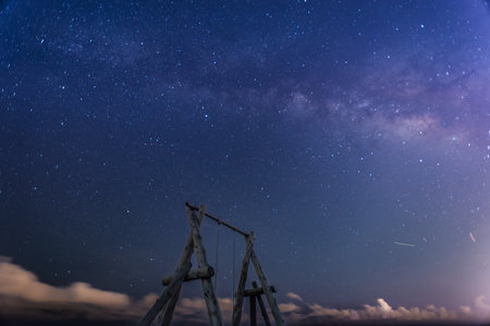 The Milky Way stretches across the night sky. A solitary wooden swing set stands on a sandy beach, inviting viewers to imagine swinging under the vast, starry sky. Zhuangwei, Yilan County, Taiwan.の写真素材