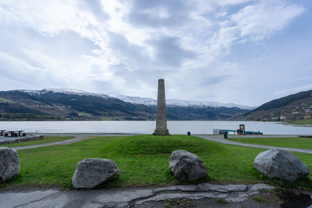 A towering stone pillar stands proudly on a grassy knoll overlooking the serene Vangsvatnet Lake in Voss, Norway. The tranquil waters reflect the surrounding mountains. Voss, Norwayの写真素材