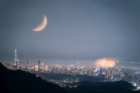 A breathtaking night view of Taipei City skyline with fireworks and a crescent moon on Qixi Festival. The romantic atmosphere is enhanced by the celestial bodies and the vibrant city lights, Taiwan.の写真素材