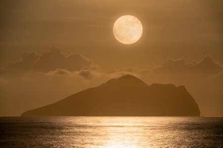 A serene night scene of the ocean with gentle waves, featuring a mesmerizing moonrise behind the iconic Turtle Island, Yilan, Taiwan. The moon gracefully dances through the clouds.の写真素材