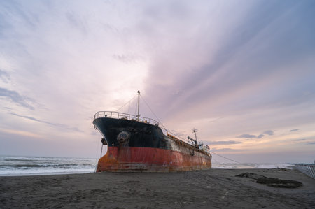 A weathered ship is silhouetted against a fiery sunset sky on the golden sands of Xigushui Beach. The contrast between the vibrant sky and the dark ship creates a dramatic seascape. Tainan, Taiwan.の写真素材