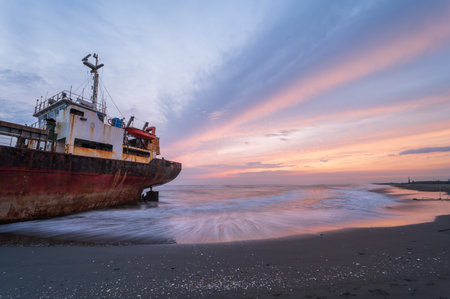 A weathered ship is silhouetted against a fiery sunset sky on the golden sands of Xigushui Beach. The contrast between the vibrant sky and the dark ship creates a dramatic seascape. Tainan, Taiwan.の写真素材