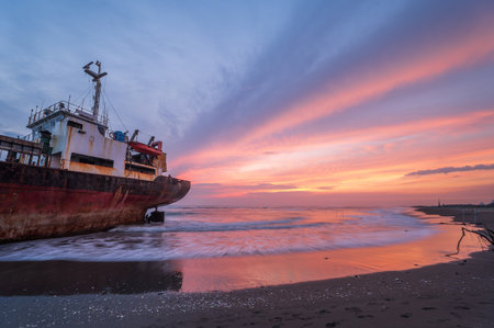 A weathered ship is silhouetted against a fiery sunset sky on the golden sands of a beach. The contrast between the vibrant sky and the dark ship creates a dramatic seascape.の写真素材