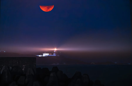 A bright red moon illuminates the night sky above a lighthouse. The lighthouse beam cuts through the...