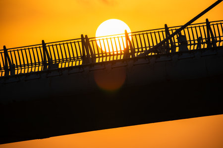 Silhouetted tourists on a bridge during a vibrant orange sunset. The sun sets behind the bridge, casting dramatic shadows.の写真素材