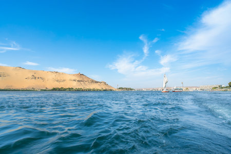 Aswan High Dam, Egypt: A felucca glides on the Nile River in spring, with sandy hills and boats along the shore under a blue sky.の写真素材
