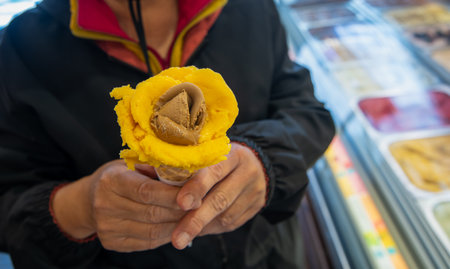 A hand holds a beautifully crafted rose-shaped gelato cone, featuring yellow mango and brown chocolate flavors, with a display case of various ice creams in the background in Lisbon, Portugal.の写真素材