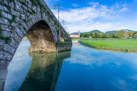 Ancient stone arch bridge over tranquil river with perfect reflections under blue sky. Historic church visible in background at Portugal.の写真素材