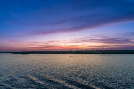 A tranquil wide shot of the Nile River at sunset near Esna Lock, Aswan, Egypt. The sky displays soft pink and purple hues, with silhouetted power towers and palm trees along the distant riverbanks.の写真素材