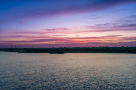 A tranquil wide shot of the Nile River at sunset near Esna Lock, Aswan, Egypt. The sky displays soft pink and purple hues, with silhouetted power towers and palm trees along the distant riverbanks.の写真素材