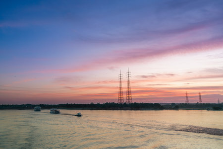 A vivid sunset with hues of pink and orange lights up the sky over the Nile River near Esna Lock, Egypt. Power transmission towers stand tall along the riverbank, framed by lush palm trees.の写真素材
