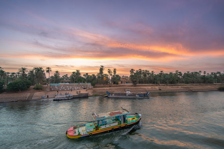 Vibrant pink and orange twilight sky above Esna Lock of The Nile, Egypt, with colorful boats on the river and lush palm trees along the bank.の写真素材