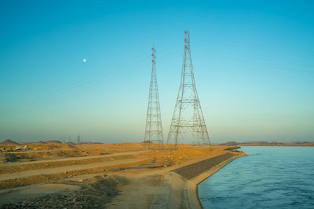 Towering power lines along the Nile River in Aswan, Egypt. The contrast between natural and man-made elements is particularly prominent as the desert terrain meets the water.の写真素材