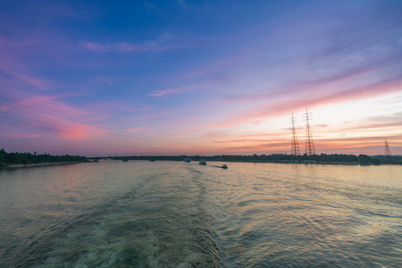 A vivid sunset with hues of pink and orange lights up the sky over the Nile River near Esna Lock, Egypt. Power transmission towers stand tall along the riverbank, framed by lush palm trees.の写真素材