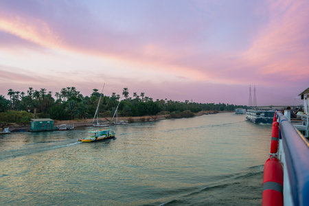 A stunning sunset with vibrant pink and orange hues colors the sky over the Nile River near Esna Lock, Aswan, Egypt. A cruise boat is visible on the tranquil waters at dusk.の写真素材