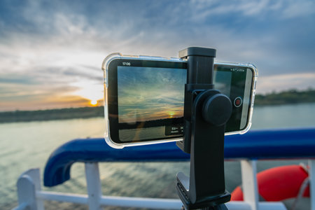 A smartphone on a tripod captures the vibrant sunset over the tranquil Nile River, as a cruise ship approaches Esna Lock in Aswan, Egypt. The scene blends technology with natural beauty.の写真素材