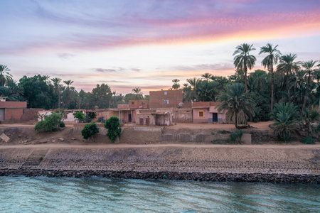 A beautiful sunset paints the sky with pink and orange hues over the Nile River near Esna Lock, Egypt. Local homes and lush palm trees line the riverbank, showcasing authentic Egyptian life.の写真素材