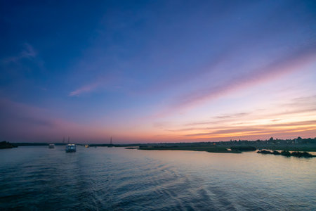 A tranquil wide shot of the Nile River at sunset near Esna Lock, Aswan, Egypt. The sky displays soft pink and purple hues, with silhouetted power towers and palm trees along the distant riverbanks.の写真素材