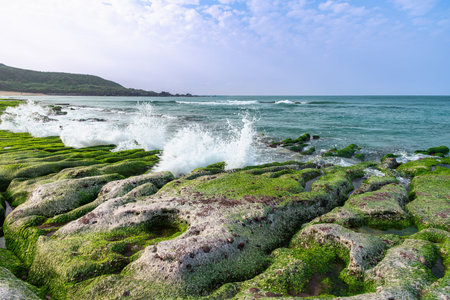 Powerful waves crash against the vibrant green algae-covered rock formations of Laomei Green Reef in...