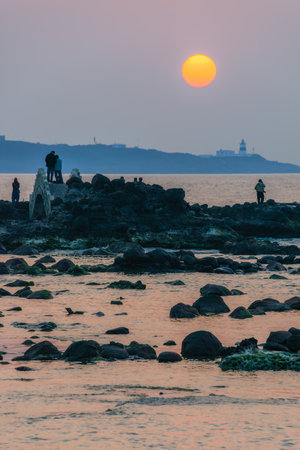Tourists gather at the rocky shore during spring sunset to admire and photograph the tranquil ocean scenery and fading sunlight over the hills. Shimen, Taiwan.の写真素材