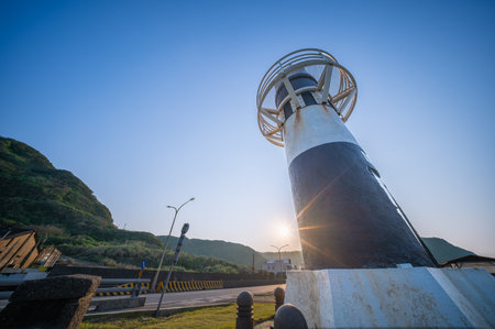 The iconic Fuguijiao Lighthouse stands tall against a clear blue sky in Shimen, New Taipei City, Taiwan, with lush green hills and sun rays.の写真素材