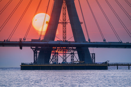 Dramatic sunset illuminating the under-construction Danshui cable bridge in New Taipei Taiwan, with the sun setting behind the towering structure over the water.の写真素材