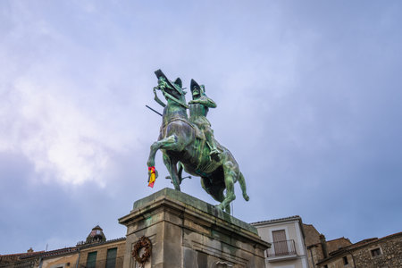 A striking bronze equestrian statue of Pizarro stands tall in Trujillo Square, Spain, under a cloudy sky. The historic town adds charm with its old buildings and warm evening lights.の写真素材