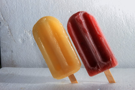 Two vibrant traditional popsicles, one yellow and one red, stand on a white textured surface,...