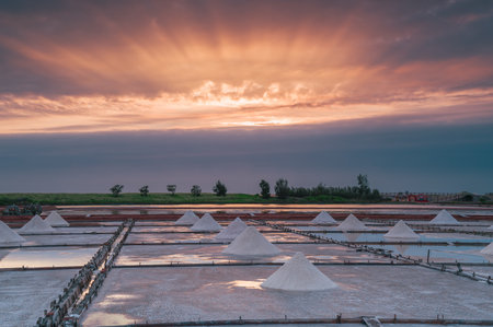 The expansive salt fields of Qigu in Tainan City, Taiwan, are bathed in the soft light of a sunset, with the sun partially obscured by dynamic clouds, creating a tranquil twilight atmosphere.の写真素材