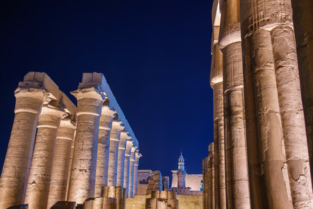 Illuminated ancient stone columns of the Temple of Luxor against a dark night sky. The minaret of the Abu Haggag Mosque is visible in the background showing two historical eras.の写真素材