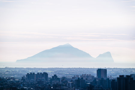 Summer dawn in Yuanshan Township, Yilan County, Taiwan, with misty Guishan Island silhouette over layered cityscape and soft pastel clouds in hazy sky.の写真素材