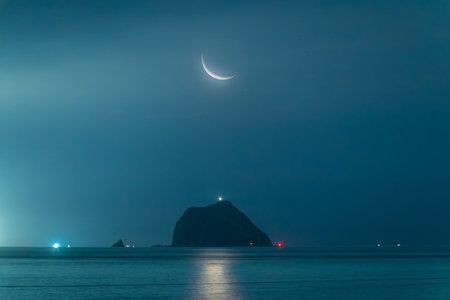 Magical night view of Keelung Islet silhouette under glowing crescent moon in Keelung, Taiwan. Calm ocean reflects golden moonlight with stars above.の写真素材