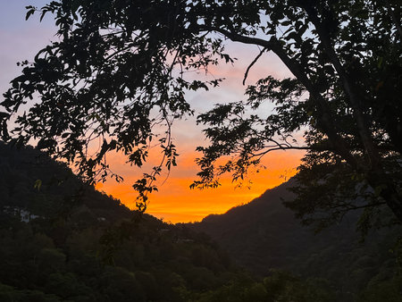 Striking orange and yellow sunrise illuminates the sky above a dark mountain valley silhouette, creating a dramatic and powerful scene.の写真素材