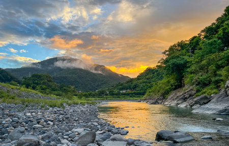 Golden sunrise light reflecting on the river water and scattered rocks in Wulai, New Taipei City, Taiwan, with low clouds and mist hanging over the lush mountains.の写真素材