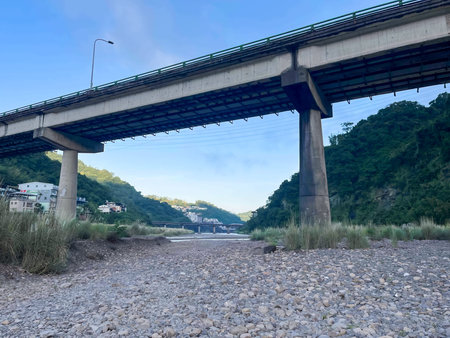 Looking up at the massive concrete structure of an elevated viaduct from the rocky riverbed. Green...