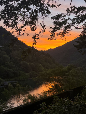 Golden sunrise light reflecting on Nanshi River, framed by overhanging tree branches and misty forested mountains in Wulai, New Taipei City, Taiwan.の写真素材