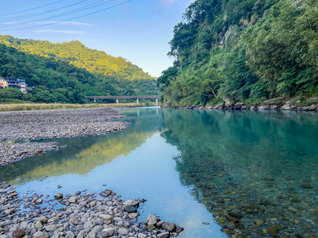 The turquoise water of the river flows past a rocky gravel bank in Wulai, New Taipei City, Taiwan. Lush green mountain slopes frame the clear stream with a distant bridge visible on a sunny morning.の写真素材