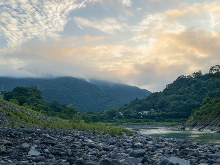 Scenic river valley view with a rocky shore and lush green mountains shrouded in mist or fog. Soft sunrise light illuminates the sky over Wulai New Taipei City Taiwan.の写真素材