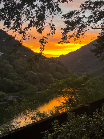 A blazing orange and yellow sunrise reflected in the river water, framed by dark tree branches and mountains in Wulai, New Taipei City, Taiwan, capturing natural intensity.の写真素材