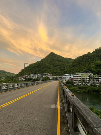 Empty bridge leading into village under a dramatic orange sunrise sky with wispy clouds, surrounded by lush green mountains, Taiwan.の写真素材