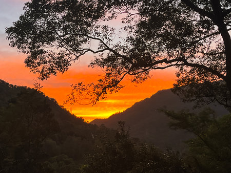 Striking orange and yellow sunrise illuminates the sky above a dark mountain valley silhouette, creating a dramatic and powerful scene.の写真素材