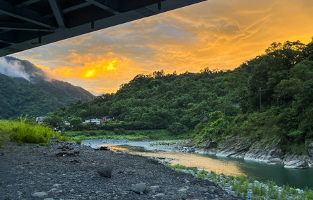 August dawn in Wulai, New Taipei City, Taiwan: orange sky, forested hills, river, and rural homes.の写真素材