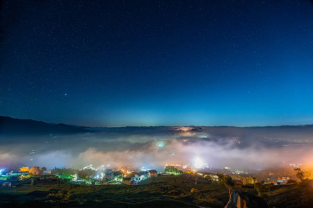 A breathtaking night view of starry sky, rolling clouds, mountain ranges, and village houses surrounded by tea plantations in Chiayi, Taiwan.