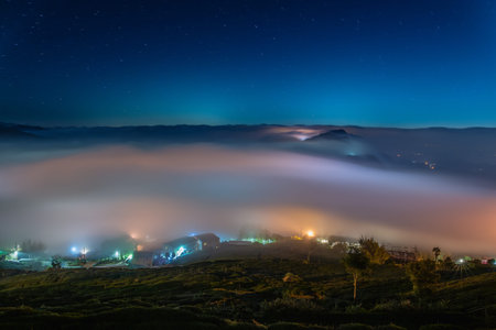 Breathtaking night view of rolling sea of clouds, distant mountain ranges, illuminated village houses, and tea plantations under a starry sky in Chiayi, Taiwan.